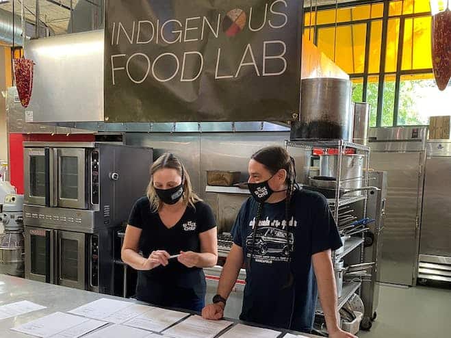 Two people standing in professional kitchen looking at papers with banner that reads "Indigenous Food Lab"